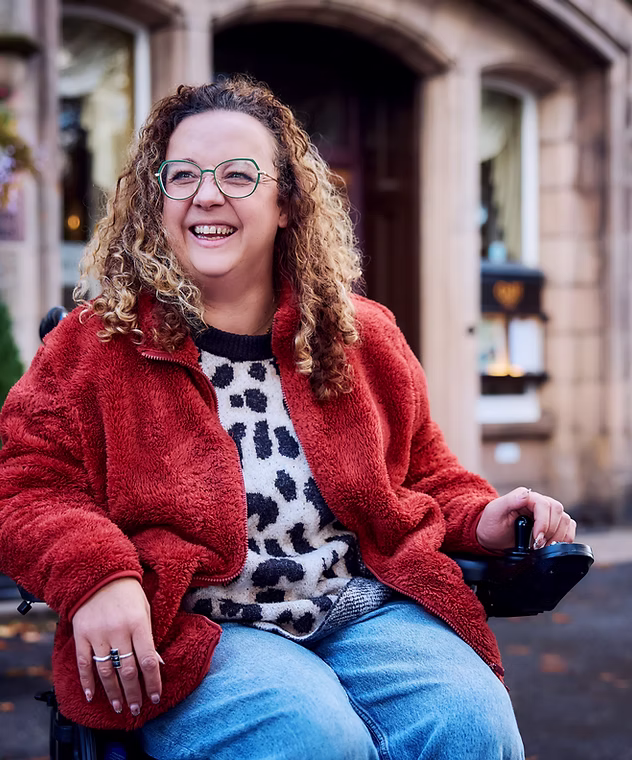 Carrie-Ann Lightley, a woman with curly hair and glasses, smiles while seated in a wheelchair outdoors in front of a building entrance. She wears a red jacket over a patterned sweater and appears relaxed and confident.